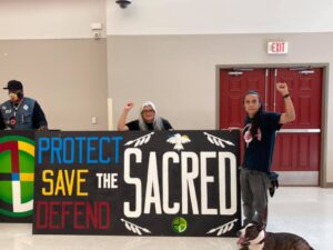 Two Indigenous men hold a sign saying, "Protect, Save, Defend the Sacred."