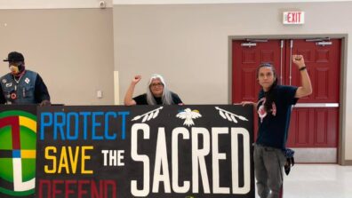 Two Indigenous men hold a sign saying, "Protect, Save, Defend the Sacred."