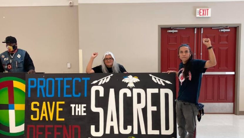 Two Indigenous men hold a sign saying, "Protect, Save, Defend the Sacred."