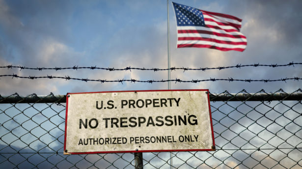 Sign on fence reading, “U.S. property No Trespassing (Authorized Personnel Only)”; American flag in the background