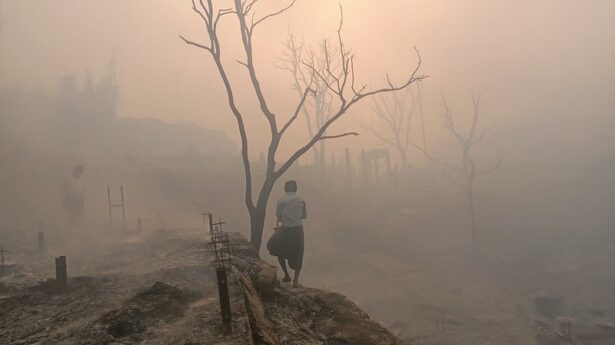 Image description: A pink mist covers the burnt remains of a Rohingya camp. Even the trees stand leafless, as if in mourning. Two people can be seen against the fading light in a landscape that is unsettling, mixing beauty with sadness. March 11, 2023