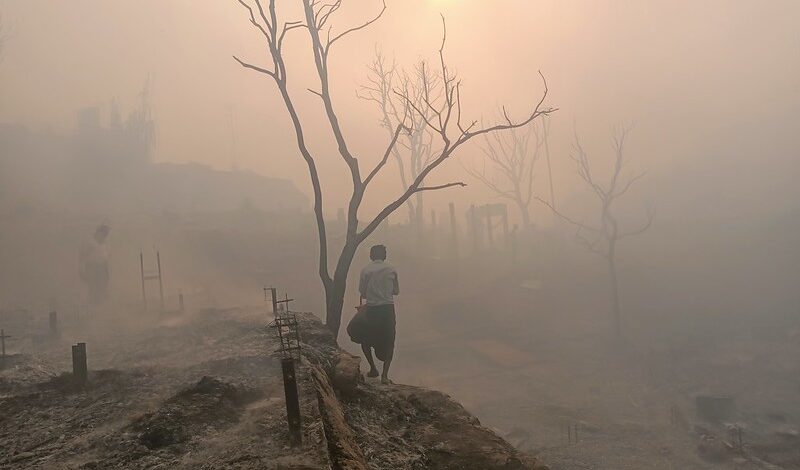 Image description: A pink mist covers the burnt remains of a Rohingya camp. Even the trees stand leafless, as if in mourning. Two people can be seen against the fading light in a landscape that is unsettling, mixing beauty with sadness. March 11, 2023