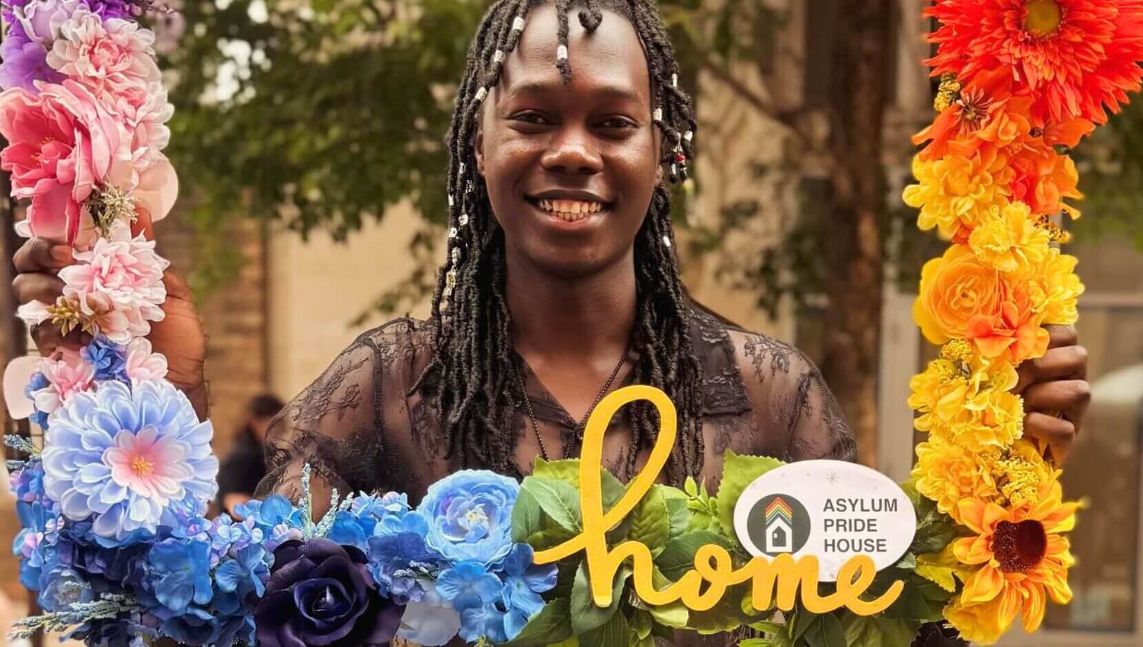 A person smiles while holding a colorful flower frame with "home" and a logo reading "Asylum Pride House." The scene exudes warmth and joy.