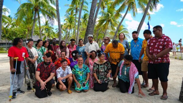 Pacific Rising 25 collective in colorful attire pose joyfully on a sandy beach. Palm trees and blue sky in the background create a tropical vibe.