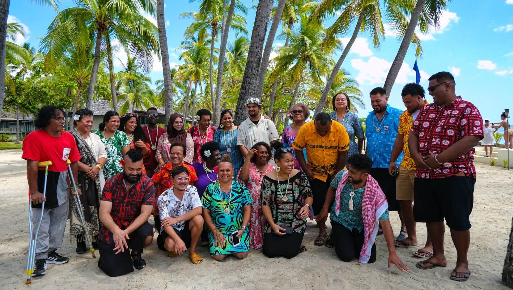 Pacific Rising 25 collective in colorful attire pose joyfully on a sandy beach. Palm trees and blue sky in the background create a tropical vibe.