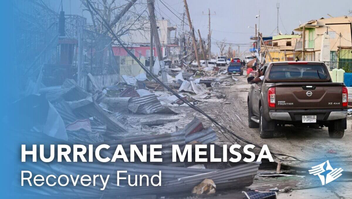 Street littered with debris from damaged buildings after Hurricane Melissa, with a truck driving through. Text overlay: "Hurricane Melissa Recovery Fund."