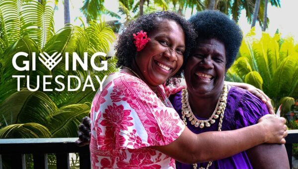 Two women Pacific Rising members embrace and smile warmly on a tropical day. One wears a pink floral dress, the other a purple dress with a necklace. "Giving Tuesday" text is visible.