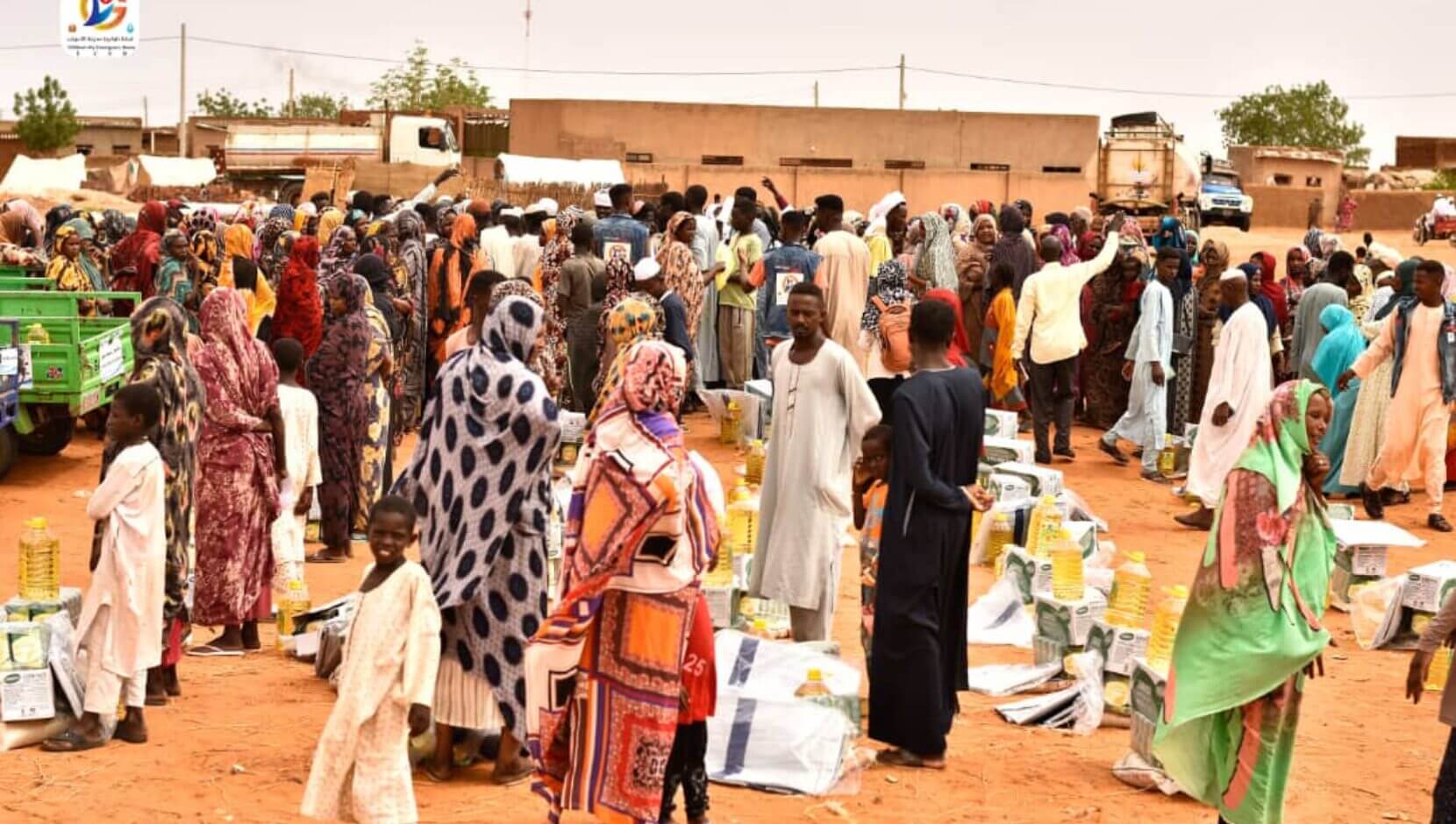 In Sudan, a large group of people, including women and children, gather outdoors among distributed supplies such as bottled oil and packaged goods.